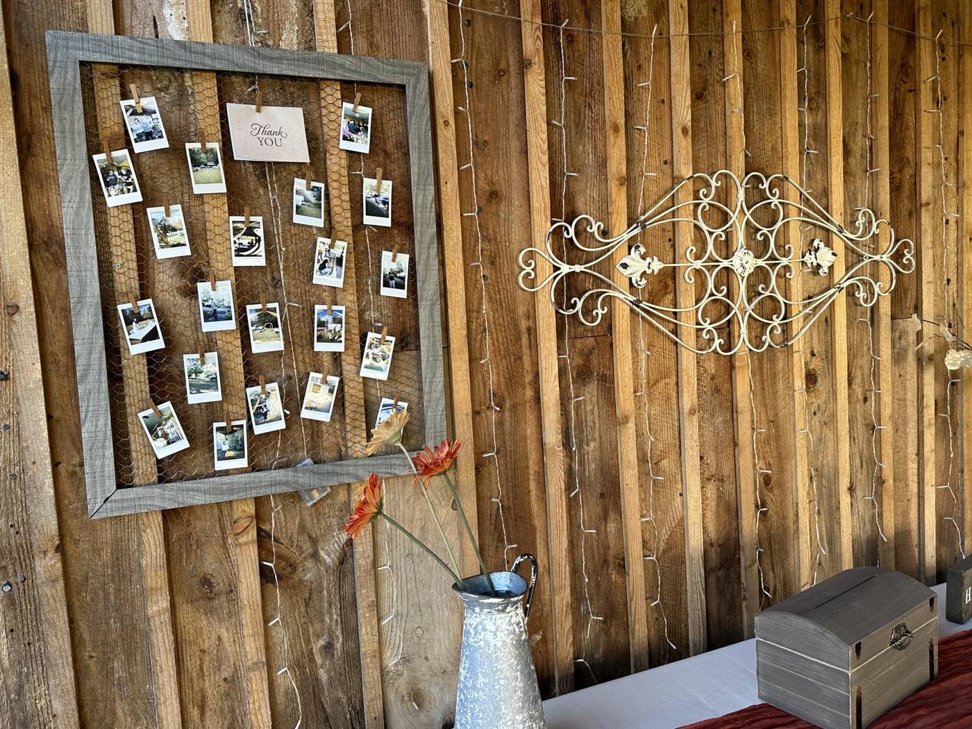 Rustic wood frame with chicken wire and clothespinned instant photos around a Thank YOU card, hanging on a barn wall beside string lights, iron scrollwork and orange gerberas in a tin pitcher