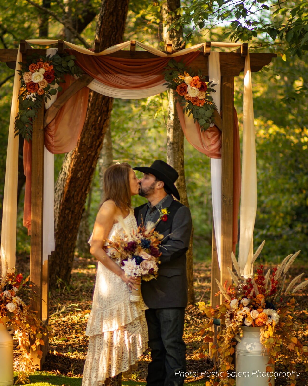 Bride and groom standing in front of the wooden ceremony arch with rust drapes and fall flowers at Riverside Farm Venue
