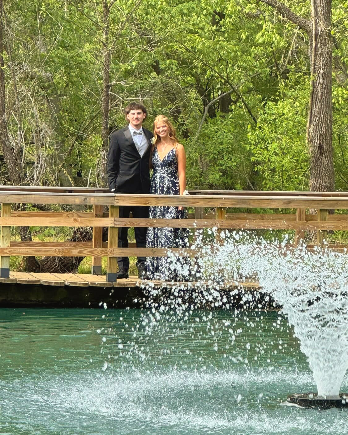 A couple dressed up on the wooden footbridge over the turquoise pond, with the fountain spraying in front, at Riverside Farm Venue