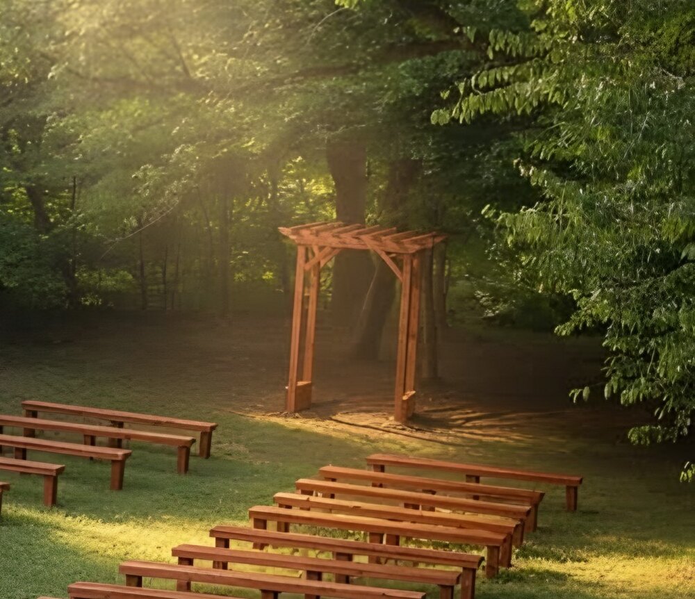 Rows of wooden benches facing the wooden ceremony arch in the sunlit woods at Riverside Farm Venue